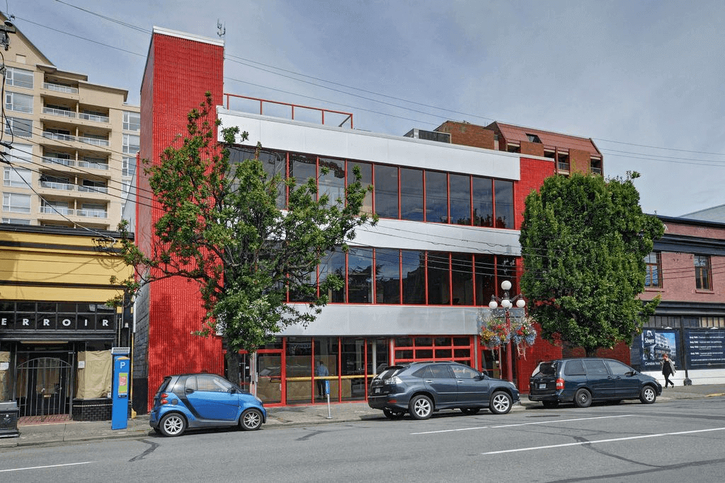 Photo prise de la rue d’un bâtiment rouge et gris et trois voitures garées devant.